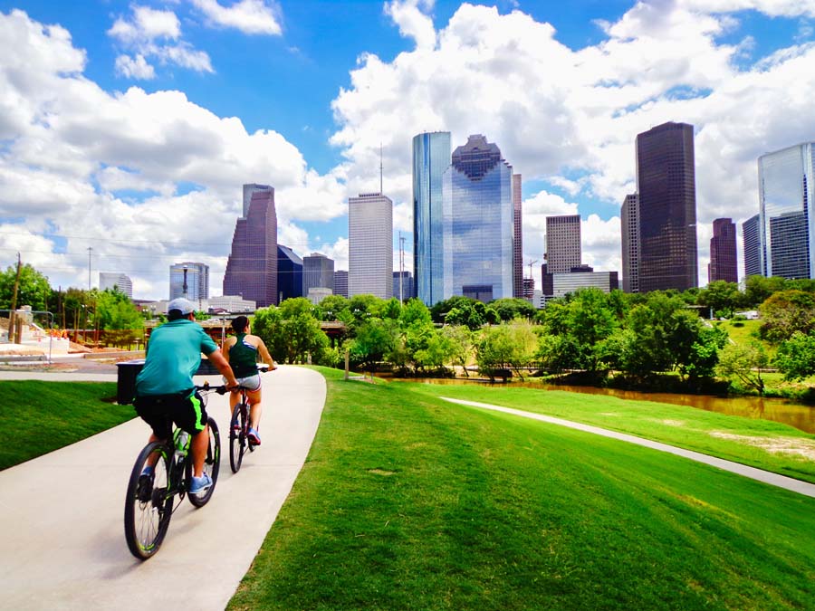 Cyclists riding along a trail in a park with a clear view of the Houston skyline in the background.