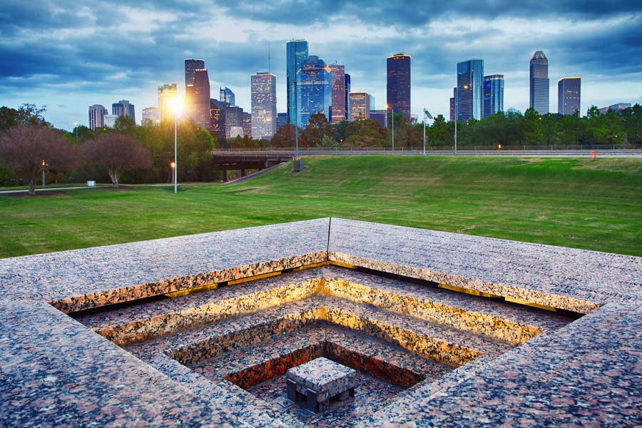 Houston cityscape seen from a park, with a monument foreground and high-rise buildings against a moody sky.