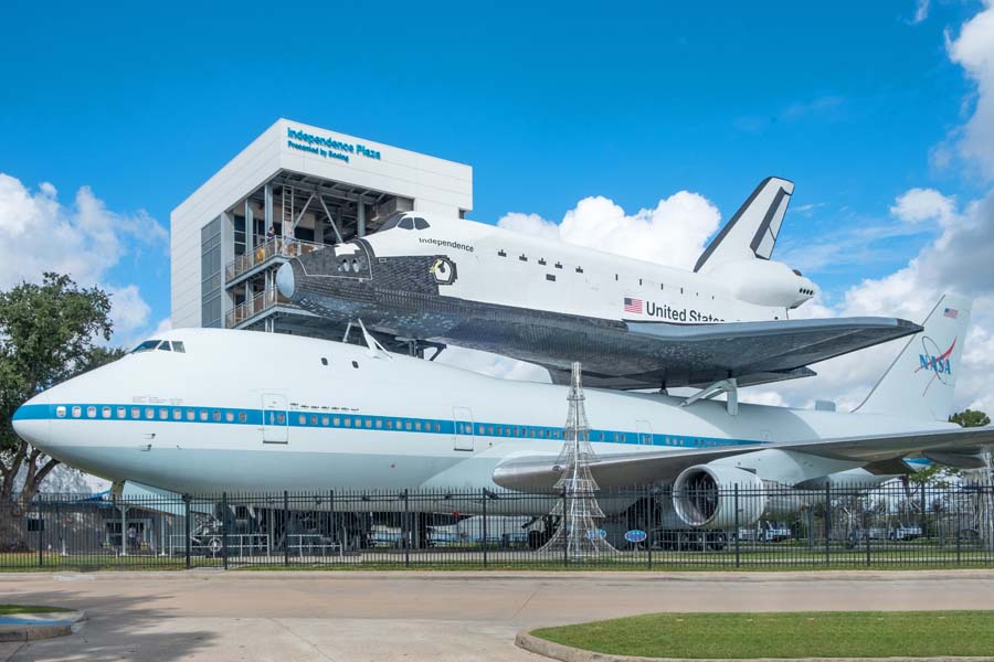 NASA's Space Shuttle replica mounted on top of a 747 aircraft at Space Center Houston, with Independence Plaza in the background.